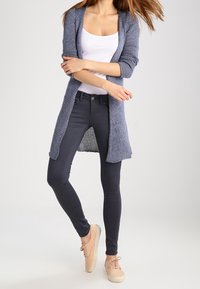 Dlouhý otevřený kardigan v námořnicky modré pletenině, nošený přes bílý nátělník a kombinovaný s tmavými úzkými džíny a světlými teniskami.