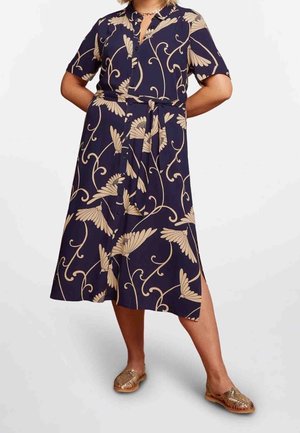 Mujer con vestido midi azul marino con estampado floral beige, mangas cortas, cinturón en la cintura y zapatos planos dorados tejidos.