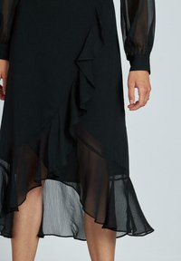 Vestido negro con volantes en capas, que presenta un dobladillo inferior transparente. Mangas largas y transparentes con puños ajustados. Textura suave y tela ligera.