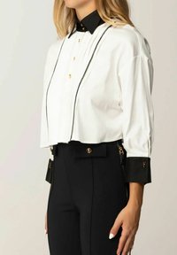 Camicia bianca crop con colletto nero, bottoni dorati e dettagli neri ai polsini; presenta una finitura strutturata e un design definito.