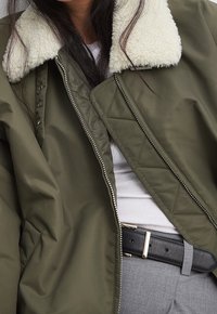 Chaqueta bomber verde oliva con cuello forrado de piel blanca, cierre de cremallera y detalles acolchados. Combinada con una camiseta blanca y pantalones grises.