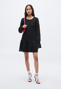 Μαύρο φόρεμα από δαντέλα με μακριά μανίκια και φαρδιά φάσα στο τελείωμα. Το μοντέλο φοράει κόκκινη τσάντα με λουράκι, λευκές κάλτσες και κόκκινα σανδάλια με τακούνι. Απλό υφασμάτινο μοτίβο.