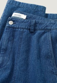 Blue denim jeans made from organic cotton, featuring a visible tag, metal button, and stitched pocket detail. Smooth texture and classic design.