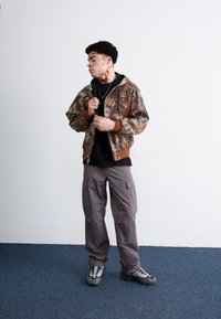 Camo bomberjack met bruine accenten, zwart hoodie shirt, grijze cargobroek en grijze sneakers. Textuurstof en relaxte pasvorm.