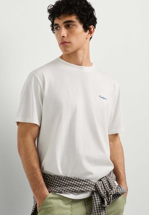 Jonge man met krullend zwart haar, dragend een wit T-shirt en groene broek met een geruit overhemd om zijn middel gebonden.