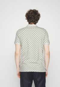 Homme aux cheveux bouclés portant un polo gris clair à manches courtes et motifs, avec un pantalon foncé, vu de dos sur un fond uni.