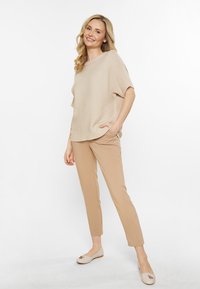Krémový pletený top s širokými rukávy, spojený s hnědými slim-fit kalhotami. Modelka má na sobě světlé krémové baleríny se zdobným akcentem u špičky.
