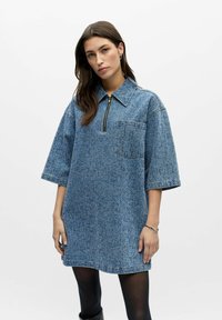 Robe surdimensionnée en jean bleu clair à manches courtes, avec col zippé, poche poitrine unique et finition texturée. Détails de couture subtils.