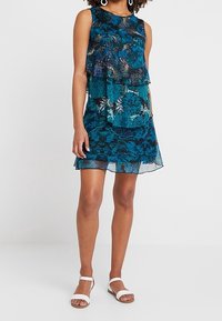 Vestido en color azul oscuro con patrones florales, capa transparente y tejido ligero. Combinado con sandalias blancas.