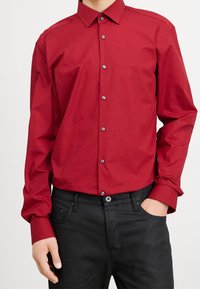 Rotes Hemd zum Zuknöpfen mit klassischem Kragen und langen Ärmeln, mit einer gleichmäßigen Textur und kontrastierenden dunklen Knöpfen, kombiniert mit schwarzer Jeans.