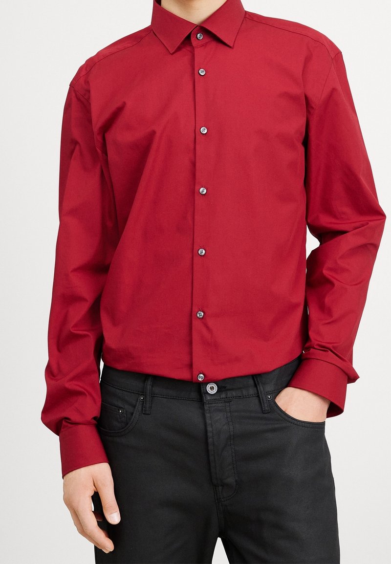 Rotes Hemd zum Zuknöpfen mit klassischem Kragen und langen Ärmeln, mit einer gleichmäßigen Textur und kontrastierenden dunklen Knöpfen, kombiniert mit schwarzer Jeans.