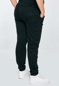 Persoon in zwarte joggingsbroek en witte sneakers, staand met handen in de zakken tegen een effen lichte achtergrond.