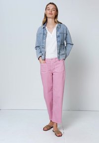 Femme debout les yeux fermés, portant une veste en jean claire, une chemise blanche, un pantalon rose avec de grandes poches et des sandales marron sur fond blanc.