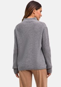 Maglione grigio lavorato a maglia con collo alto, texture a coste e maniche lunghe, abbinato a pantaloni marrone chiaro, mostrato da dietro.
