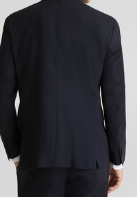 Giacca di suit blu scuro realizzata in tessuto morbido. Presenta un singolo spacco sul retro, con un sottile motivo a quadri visibile sui polsini.
