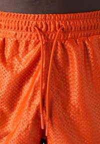 Shorts arancioni in rete con cintura elasticizzata, dotati di cordino e piccole perforazioni per la traspirabilità. Texture liscia e design leggero.