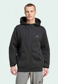 Felpa con cappuccio nera con cerniera, realizzata in materiale morbido, con tasca frontale, cappuccio regolabile e logo Adidas in grigio scuro sul lato sinistro del petto.