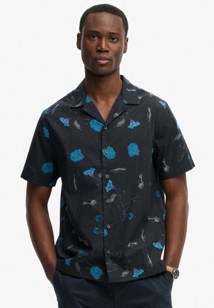 Mężczyzna ubrany w czarną koszulę na guziki z krótkim rękawem, ozdobioną niebiesko-fioletowymi kwiatowymi wzorami, z rękami w kieszeniach, na neutralnym tle.