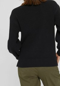 Czarny, dziergany sweter o teksturowanej powierzchni, z prążkowanym dekoltem i mankietami. Krój jest luźny, zestawiony z oliwkowozielonymi spodniami.