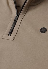 Vêtement beige à demi-fermeture éclair avec un curseur noir marqué "Street One Men" et un patch rond noir avec un logo carré sur la poitrine.