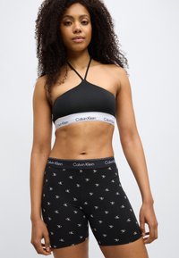 Γυναίκα που φοράει μαύρο κοντό τοπ halter Calvin Klein και μαύρο σορτς με το λογότυπο Calvin Klein, στέκεται μπροστά σε λευκό φόντο.