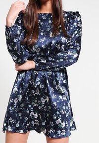 Robe florale bleu marine en satin avec des manches bouffantes, une taille froncée et une jupe évasée ; présente des motifs floraux blancs et verts.