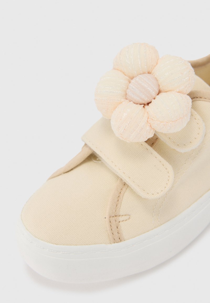 Beige Leinenschuhe mit einer weißen Gummisohle, die mit einer großen Stoffblumen-Applikation in Pastellfarben und einem Klettverschluss ausgestattet sind.