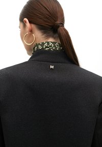 Black textured blazer with a small embroidered ‘M’ on the back. The model wears a green polka-dot scarf and large gold hoop earrings.