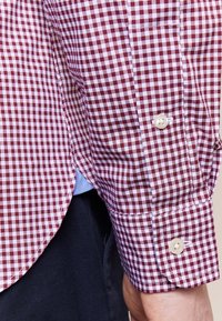 Rutig skjorta i röd och vit, av bomullstyg, med knäppta ärmslut, rundad nederkant, och kontrasterande blått innerfoder synligt längst ner.