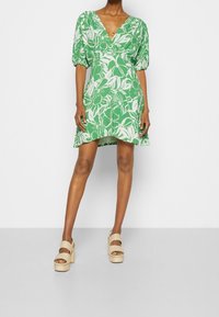 Zelené květinové šaty s výstřihem do V, nabíranými rukávy a vrstevnatým lemem. Modelka má na sobě béžové platformové sandály se dvěma řemínky kolem kotníku.