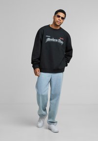 Hanorac negru cu grafică "Members Only", asociat cu blugi albăstrii deschiși și adidași albi. Modelul poartă ochelari de soare. Stil casual, croială lejeră.