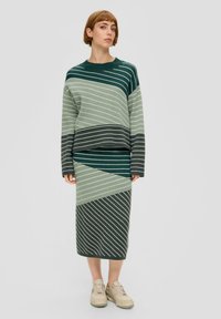 Gebreide jurk met groene, grijze en witte diagonale strepen, met een losse pasvorm en lange mouwen, gecombineerd met beige sneakers.