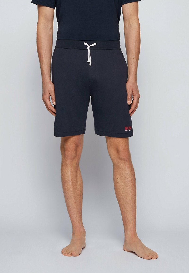 Homme portant un short bleu marine longueur genou avec cordon blanc et logo rouge "BOSS" sur la jambe gauche, debout pieds nus devant un fond gris.