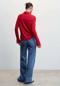 Polo shirt rouge à manches longues avec un col plat, associé à un jean en denim bleu à coupe ample. La texture douce et la coupe décontractée sont visibles.