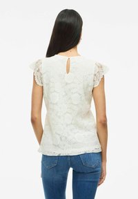 Hvid blondebluse med korte flæseærmer, der har en nøglehulslukning bagpå og komplekse blomstermønstre, parret med blå jeans.
