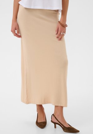 Femme portant une jupe midi beige, un haut blanc, des talons slingback marron, des bagues aux doigts et un bracelet marron au poignet droit.