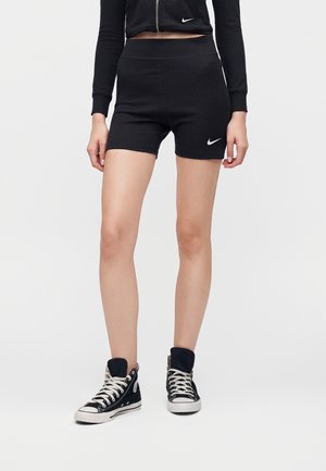 Kobieta w czarnych szortach Nike z wysokim stanem, czarnej krótkiej kurtce na suwak i czarnych wysokich trampkach z białymi sznurowadłami na jednolitym tle.