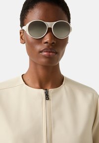 Okrągłe, jasne okulary przeciwsłoneczne z przyciemnianymi szkłami, zestawione z beżową kurtką z wysokim kołnierzem i zamkiem z przodu. Materiał o miękkiej fakturze, minimalistyczny design.