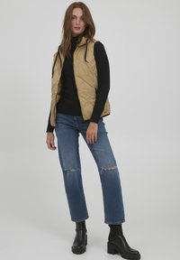 Beige gesteppte Weste mit hohem Kragen, schwarzes langärmeliges Oberteil und ausgebleichte blaue Jeans. Schwarze klobige Stiefel bis zur Knöchel runden das Outfit ab.