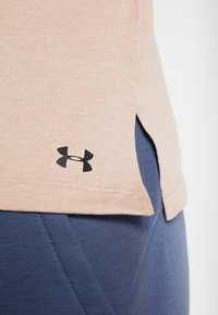 Top sportivo rosa chiaro con orlo arrotondato e fessure sui lati, con logo Under Armour nero in basso a sinistra. Abbinate a leggings blu.