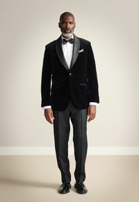 Zwarte fluwelen smokingjack met satijnen revers, witte overhemd, zwarte vlinderdas en zwarte pantalon; glanzende zwarte schoenen.