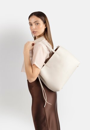 Donna con top beige a maniche corte e gonna marrone porta una grande borsa bianca sulla spalla, guardando verso la macchina fotografica con sfondo semplice.