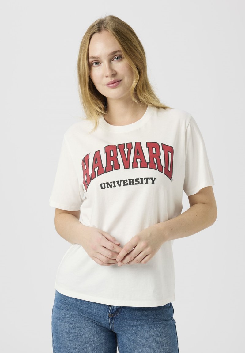 Cremefarbenes Baumwoll-T-Shirt mit auffälligem rotem "HARVARD"-Schriftzug und schwarzem "UNIVERSITY" darunter. Kurzärmelig und mit lockerer Passform. Kombiniert mit blauen Jeans.