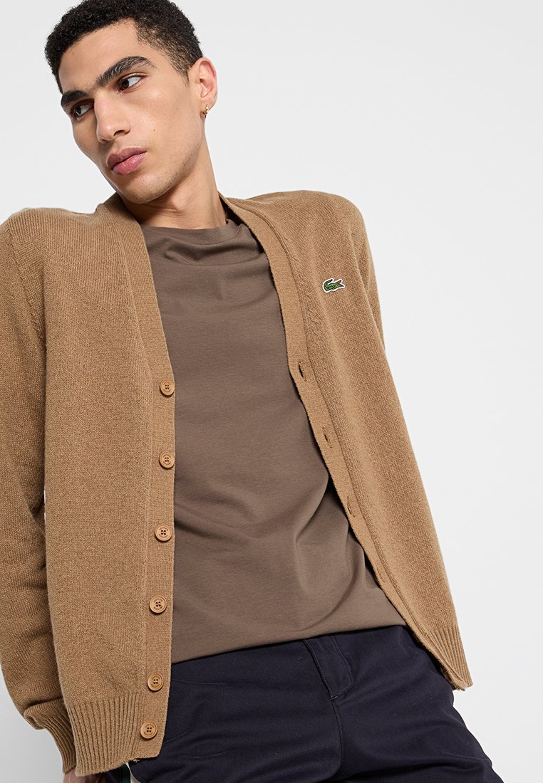 Cardigan tricoté marron avec des boutons, présentant un petit logo vert. Porté par-dessus un t-shirt marron, associé à un pantalon sombre. Texture lisse.