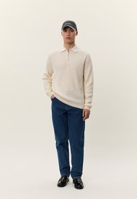 Mann trägt cremefarbenen Pullover mit Reißverschluss am Kragen, dunkelblaue Jeans, schwarze Schuhe und dunkelgraue Baseballkappe, steht mit einer Hand in der Tasche.