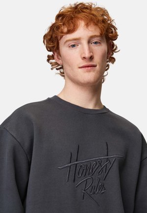 Mørkegrå sweatshirt med rund halsudskæring og raglanærmer. Har struktureret broderi af sætningen "Ærlighed Regler" i kontrasterende tråd.