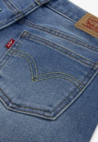 Blue denim jeans featuring a back pocket with a yellow heart-shaped stitch detail and a brown leather patch with brand logo.