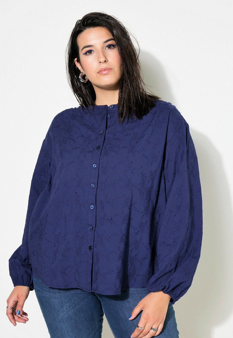 Camicia azzurra con bottoni, motivo floreale testurizzato, colletto arrotondato e maniche lunghe e voluminose. Abbinata a jeans blu.
