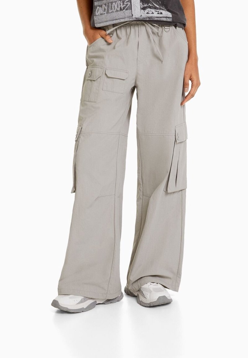 Šedé cargo kalhoty s volnějším střihem, které mají více kapes, elastický pas a lehký materiál. Textura vypadá hladce.