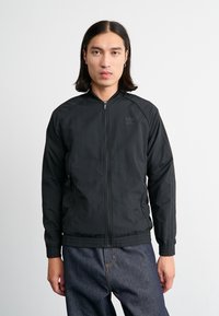Juodas zip-up švarkas, pagamintas iš lengvos medžiagos, turintis stovinčią apykaklę, elastingas rankogalius ir siuvinėtą logotipą kairėje krūtinės pusėje.